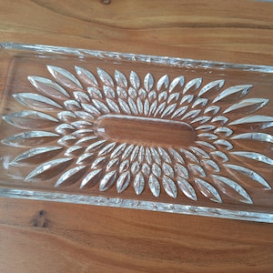 May include: A clear glass serving tray with a decorative, sunburst-like pattern. The tray is rectangular and has a slightly raised edge.