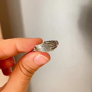 May include: A silver ring with a detailed wing design. The ring is held by a finger.