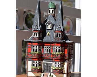 May include: A red and black ceramic model of a building with a clock tower. The building has a pointed roof and decorative details. The model is a traditional German style.