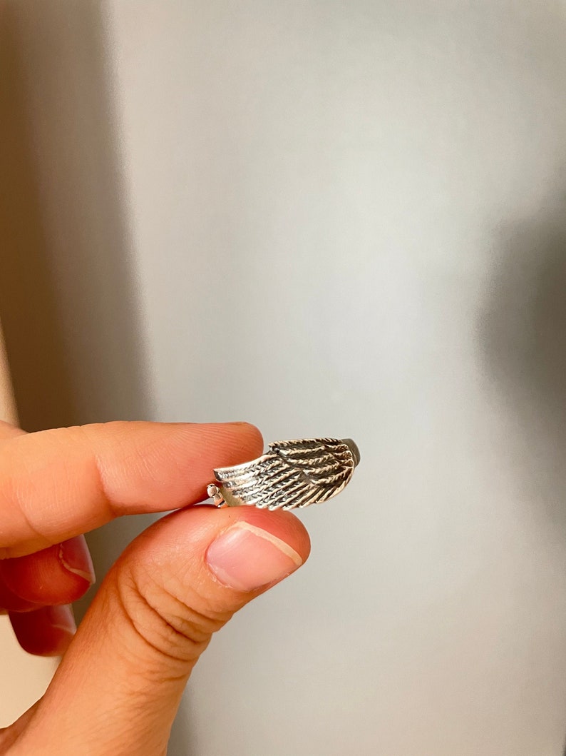 May include: A silver ring with a detailed wing design. The ring is held by a finger.