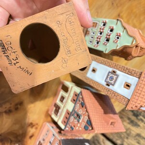 May include: A set of five miniature ceramic houses with a terracotta base. The houses are decorated with white and green paint and have intricate details. The base is labeled "Ratheniburg ob der Tauber" and "Goth 31 Mini" in black lettering.