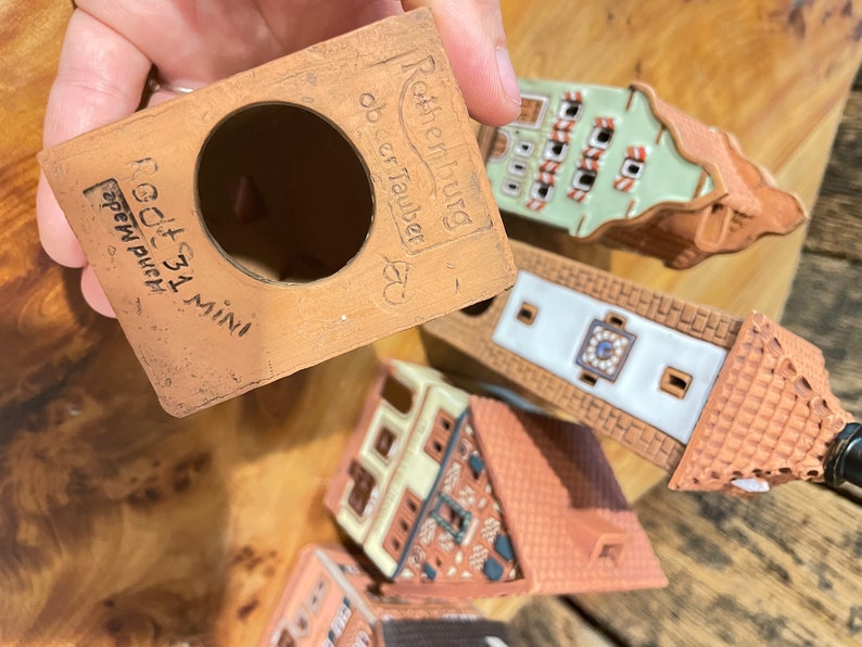 May include: A set of five miniature ceramic houses with a terracotta base. The houses are decorated with white and green paint and have intricate details. The base is labeled "Ratheniburg ob der Tauber" and "Goth 31 Mini" in black lettering.
