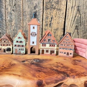 May include: Five miniature ceramic houses with painted details, including windows, doors, and roofs. The houses are arranged in a row on a wooden surface. The houses are painted in shades of brown, green, and white. The text on the houses reads "Zur Post", "Gasthaus", "Hofbrau", and "Anklerschloss".
