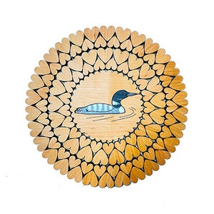 以下が含まれることがあります： 湖で泳ぐ loon を囲むハートのデザインが施された丸い木製の壁飾り。loon は黒と白で、背中に青いストライプがあります。