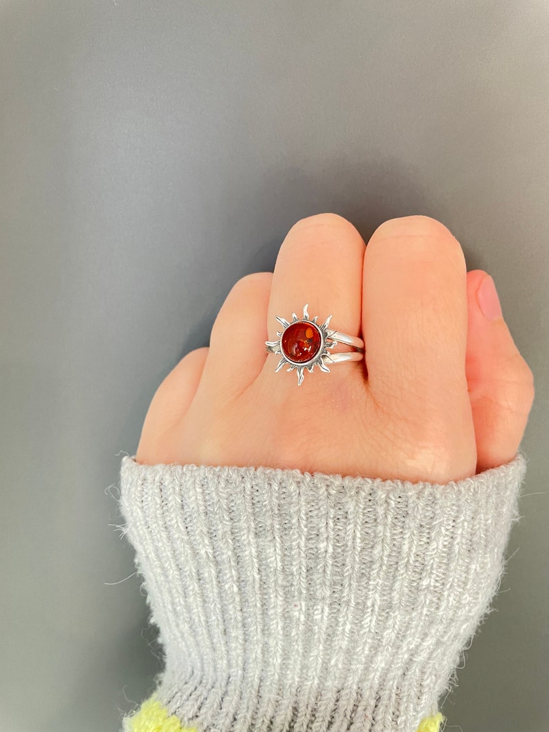 K&ouml;nnte beinhalten: Ein silberner Ring mit Sonnenmuster. Er hat einen runden, bernsteinfarbenen Stein in der Mitte, umgeben von strahlenf&ouml;rmigen Zacken. Der Ring wird an einem Finger getragen und zeigt seine komplizierten Details und seine warme Farbe.