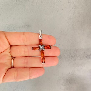 Puede incluir: Un colgante de cruz de plata con un centro de &aacute;mbar marr&oacute;n. La cruz est&aacute; decorada con peque&ntilde;os detalles plateados.