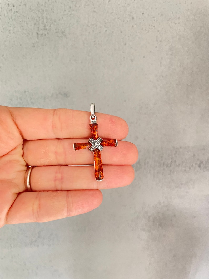 Puede incluir: Un colgante de cruz de plata con un centro de &aacute;mbar marr&oacute;n. La cruz est&aacute; decorada con peque&ntilde;os detalles plateados.