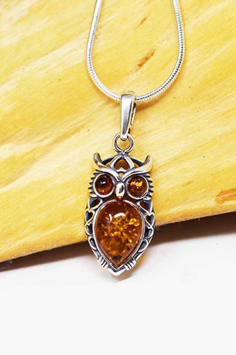May include: Silver owl pendant necklace featuring a teardrop-shaped amber stone. The owl pendant has detailed features, including eyes and a textured body. The necklace is on a silver chain, set against a light brown wooden surface.