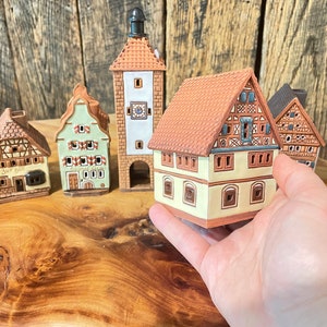 May include: Five miniature ceramic houses with intricate details, including windows, doors, and roofs. The houses are painted in various colors, including brown, green, and white. The houses are arranged on a wooden surface.