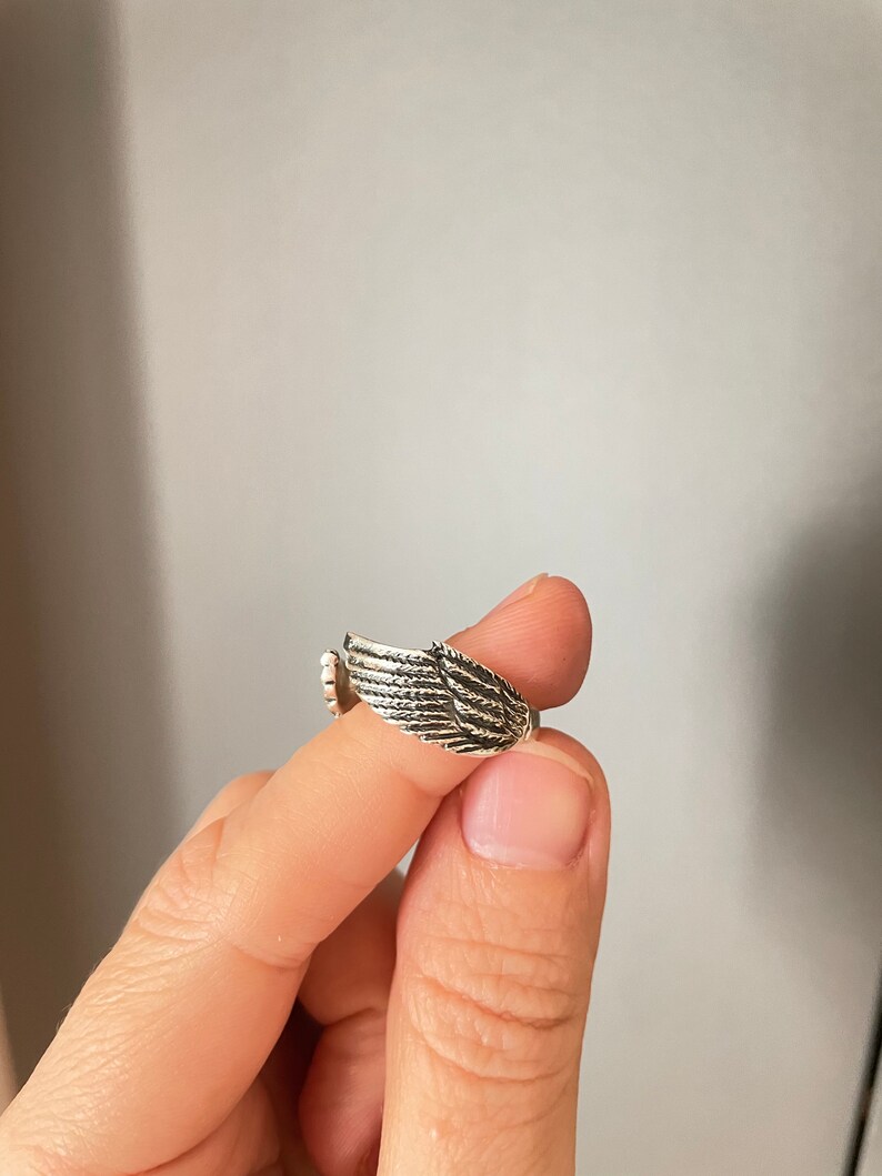 May include: A silver ring with a wing design. The ring is on a finger.