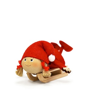 May include: A wooden figurine of a girl wearing a red hat and a red dress, riding a wooden sled. The girl has a smiling face and blonde hair.