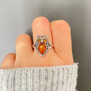 May include: A silver owl ring with amber-coloured eyes and a pear-shaped amber body. The ring is worn on a finger, against a grey background. The owl design has intricate details.