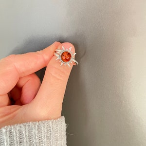 K&ouml;nnte beinhalten: Ein silberner Ring mit einem Sonnenmotiv. Der Ring hat einen zentralen, bernsteinfarbenen Stein, der von einem Sonnenstrahlenmuster umgeben ist. Der Ring wird von einer Hand gehalten, wodurch das Design und die Farbe des Rings hervorgehoben werden.