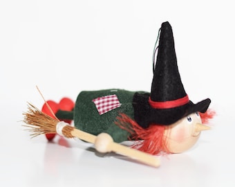Può includere: Una bambola di strega di legno con capelli rossi, un cappello nero e un vestito verde sta volando su una scopa. La bambola ha un viso dipinto con guance rosate e un ampio sorriso.