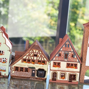 May include: Four miniature ceramic houses with intricate details, including windows, doors, and roofs. The houses are painted in various colors, including green, brown, and white. The houses are arranged in a row on a glass surface. The houses have signs that read "Altstadt Garten", "Mittlere Strasse", "B&auml;ckerei Braun", and "Rathaus".