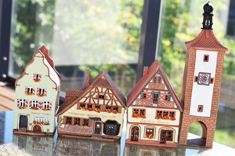 May include: Four miniature ceramic houses with intricate details, including windows, doors, and roofs. The houses are painted in various colors, including green, brown, and white. The houses are arranged in a row on a glass surface. The houses have signs that read "Altstadt Garten", "Mittlere Strasse", "B&auml;ckerei Braun", and "Rathaus".