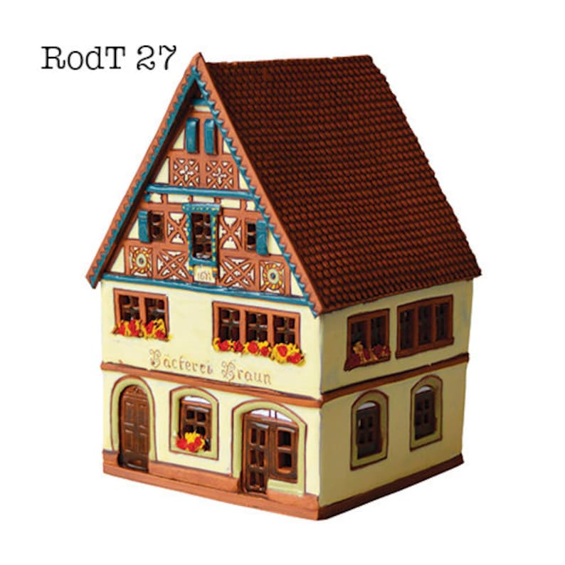 May include: A small, yellow, ceramic house with a brown roof and a sign that reads "Backerei Braun". The house has a detailed facade with blue and white trim and a decorative wooden design.