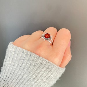 K&ouml;nnte beinhalten: Ein silberner Ring mit einem Sonnenmuster und einem runden, rot-orangen Edelstein in der Mitte. Der Ring wird an einer Hand getragen, wodurch die detaillierte Gestaltung und die Farbe des Steins hervorgehoben werden. Das Design des Rings erinnert an ein Himmelsmotiv.