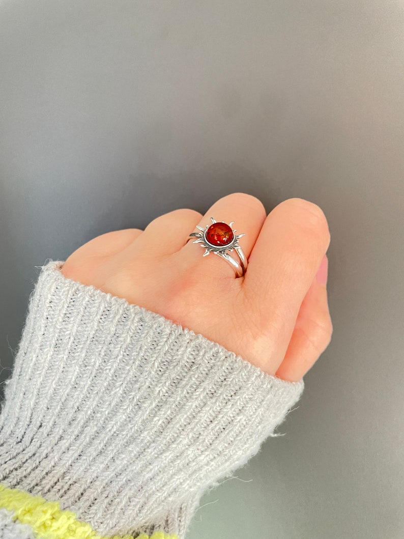 K&ouml;nnte beinhalten: Ein silberner Ring mit einem Sonnenmuster und einem runden, rot-orangen Edelstein in der Mitte. Der Ring wird an einer Hand getragen, wodurch die detaillierte Gestaltung und die Farbe des Steins hervorgehoben werden. Das Design des Rings erinnert an ein Himmelsmotiv.