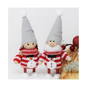 Peut inclure: Deux lutins de Noël, une femme et un homme, avec des chapeaux blancs et des pulls en tricot rayés rouge et blanc. Les lutins sont debout sur une surface en bois avec un fond brun.