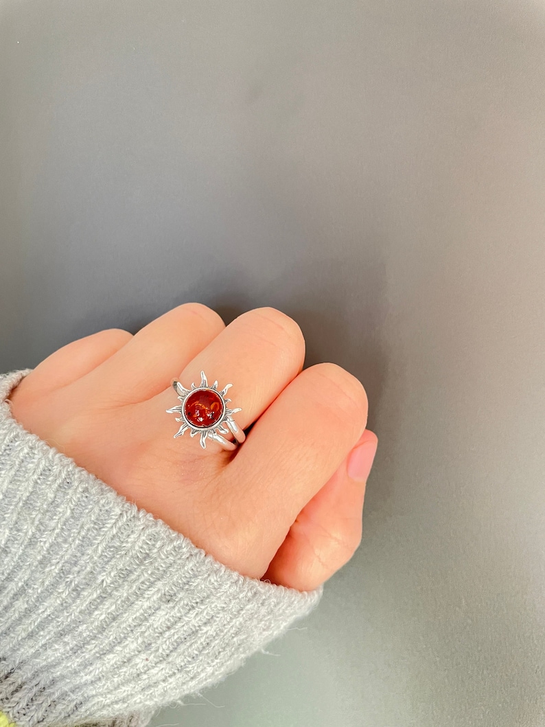 K&ouml;nnte beinhalten: Ein silberner Ring mit einem Sonnenmuster und einem zentralen, runden, rot-orangen Stein. Der Ring wird am Finger getragen und zeigt die detaillierten Sonnenstrahlen und die leuchtende Farbe des Steins.