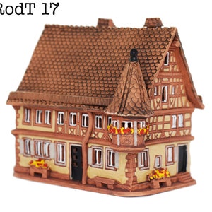 May include: A small, brown, ceramic model of a traditional German half-timbered house with a red tiled roof. The house has a balcony with red flowers and a small window. The house is decorated with white trim and has a brown door.