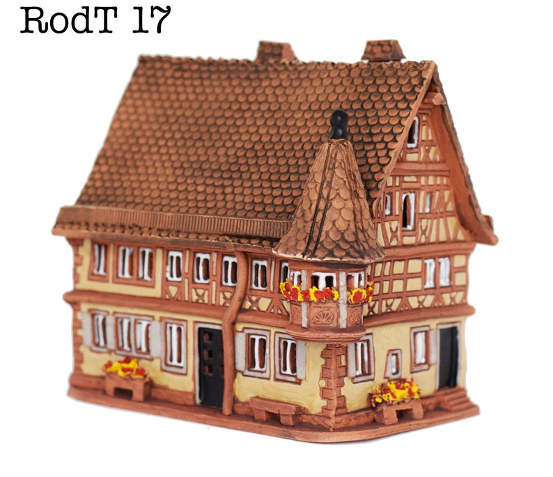 May include: A small, brown, ceramic model of a traditional German half-timbered house with a red tiled roof. The house has a balcony with red flowers and a small window. The house is decorated with white trim and has a brown door.