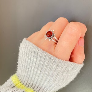 K&ouml;nnte beinhalten: Ein silberner Ring mit einem Sonnenmuster und einem zentralen, bernsteinfarbenen Stein. Das Sonnenmuster des Rings hat spitze Strahlen um den Stein herum. Der Ring wird an einem Finger getragen. Schmuck.