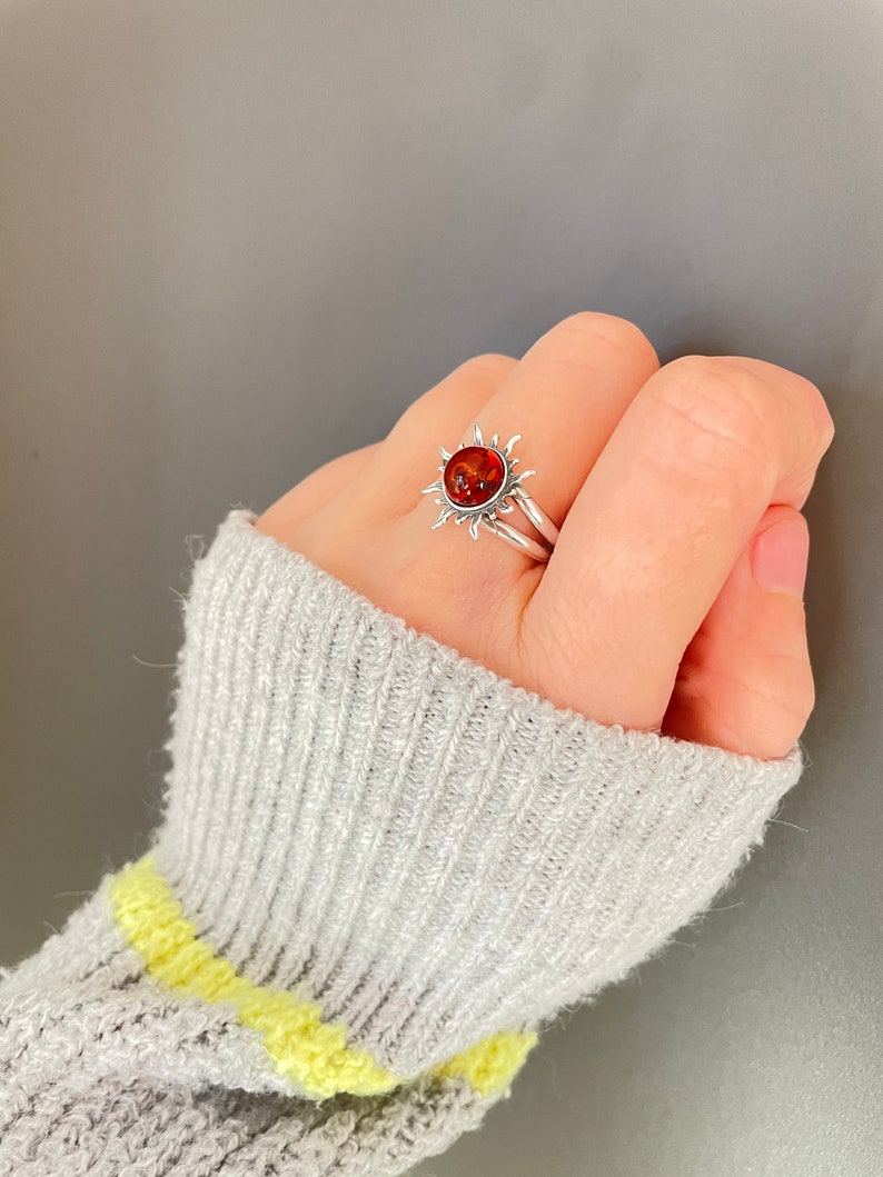 K&ouml;nnte beinhalten: Ein silberner Ring mit einem Sonnenmuster und einem zentralen, bernsteinfarbenen Stein. Das Sonnenmuster des Rings hat spitze Strahlen um den Stein herum. Der Ring wird an einem Finger getragen. Schmuck.