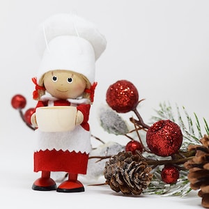 May include: A small wooden doll wearing a white chef's hat and a red dress, holding a white cup. The doll is standing in front of a snowy pine branch with red berries and a pine cone.