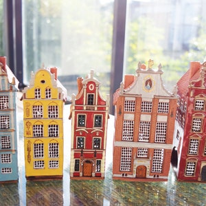 May include: Five miniature ceramic houses in different colors, including blue, yellow, red, and brown. Each house has multiple windows and a detailed facade.
