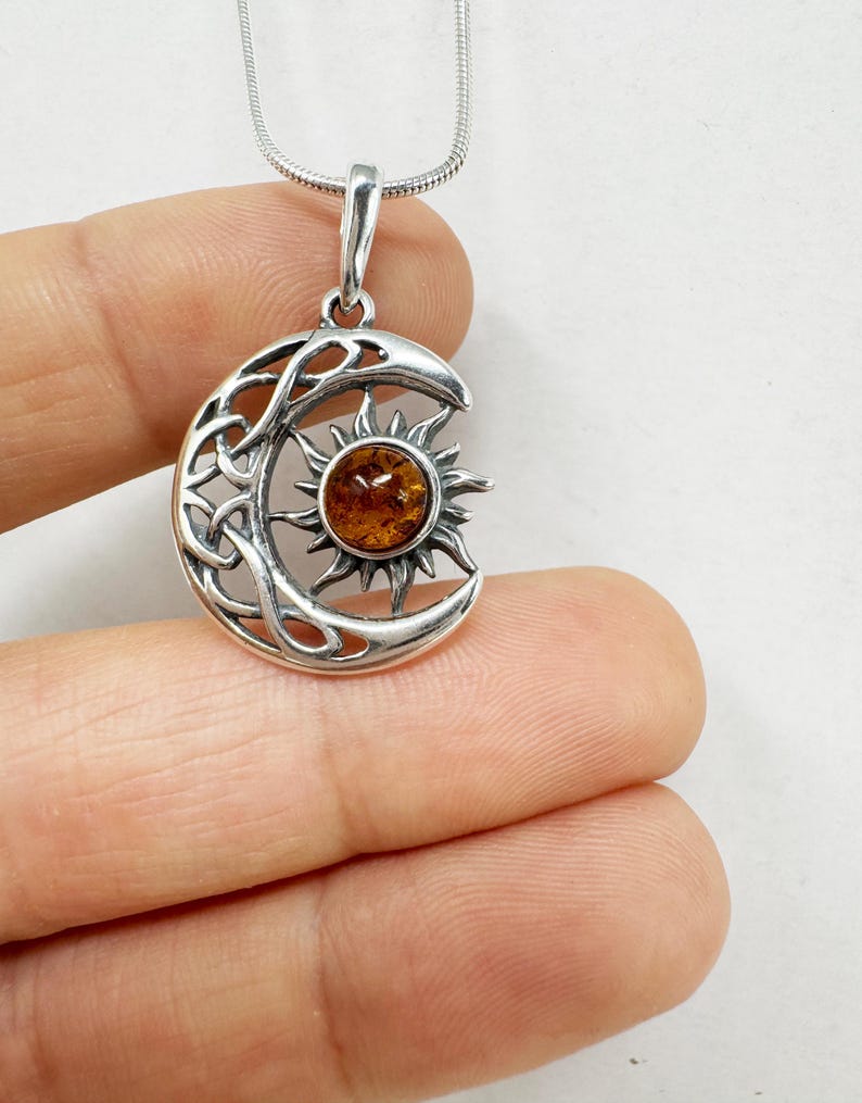 May include: A silver pendant necklace featuring a crescent moon design with Celtic knotwork and a sunburst. The sun in the center is a round amber stone. The necklace has a silver chain.