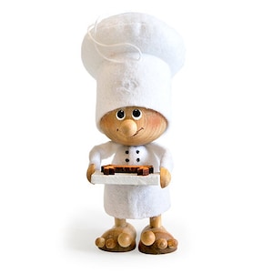 May include: A wooden figurine of a baker wearing a white chef's hat and holding a tray with a brown pastry. The baker has a smiling face with a large nose and brown eyes.