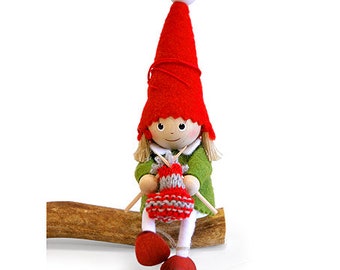 May include: A handmade doll with a red pointed hat and a white ball on top. The doll wears a green jacket and white pants, and red shoes. It is knitting a red and white item with wooden needles. The doll is sitting on a brown wooden log.