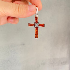 Puede incluir: Un colgante de cruz de plata con una incrustaci&oacute;n de piedra &aacute;mbar marr&oacute;n. La cruz est&aacute; detallada con peque&ntilde;os detalles plateados.