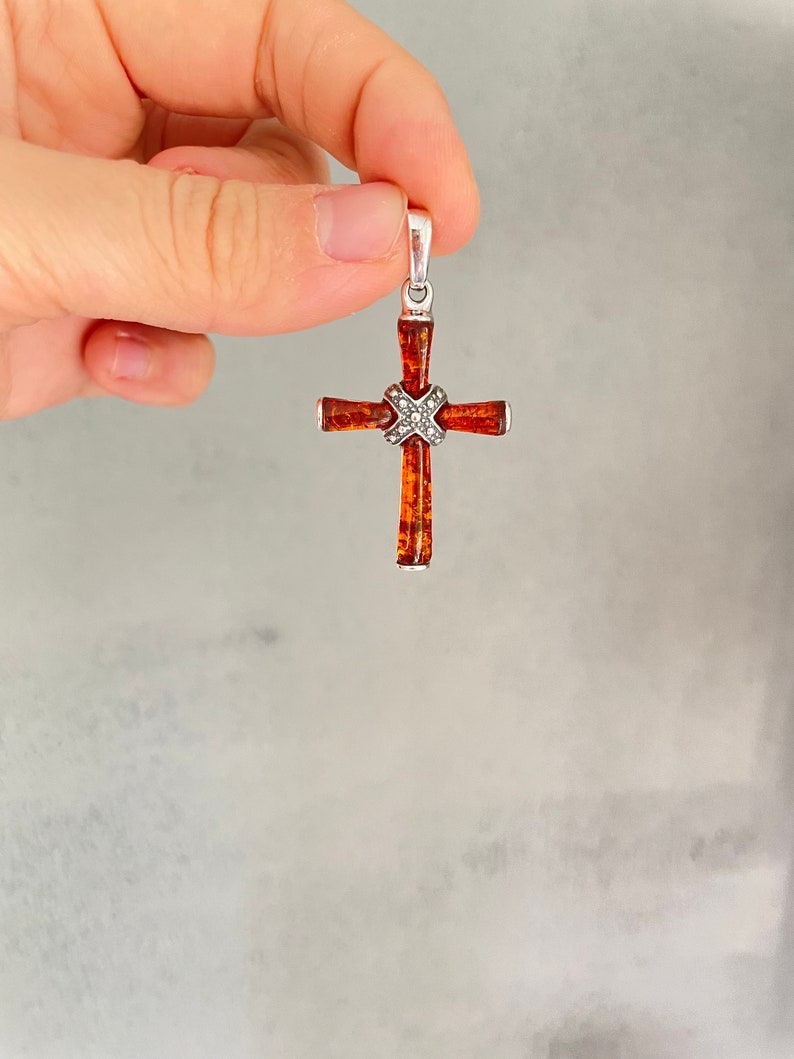 Puede incluir: Un colgante de cruz de plata con una incrustaci&oacute;n de piedra &aacute;mbar marr&oacute;n. La cruz est&aacute; detallada con peque&ntilde;os detalles plateados.
