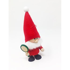May include: A red and white Christmas gnome holding a green and white tennis racket. The gnome has a white beard and a red hat with a white pom-pom.