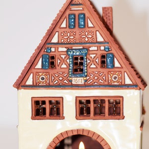 May include: A ceramic house shaped like a traditional German half-timbered house with a candle holder in the front. The house is decorated with blue and white details and the year "1617" is inscribed on the front.