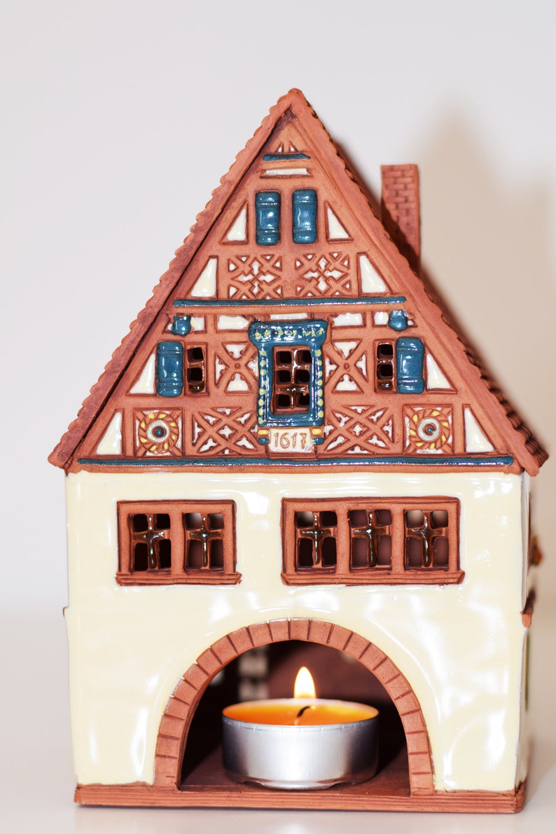 May include: A ceramic house shaped like a traditional German half-timbered house with a candle holder in the front. The house is decorated with blue and white details and the year "1617" is inscribed on the front.