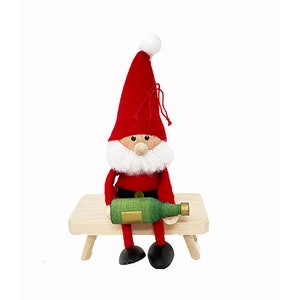 May include: A red felt gnome with a white beard and a green bottle in its hands, sitting on a wooden bench.
