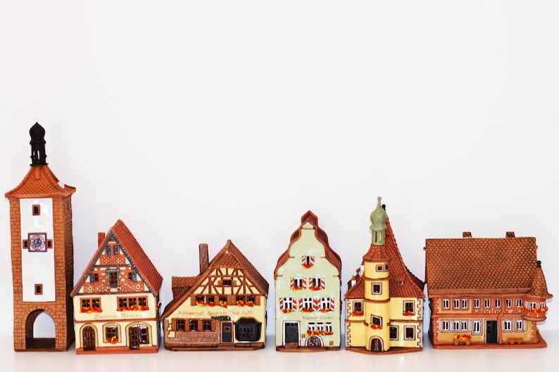 May include: A collection of six miniature ceramic houses with brown, yellow, and green paint. The houses have intricate details, including windows, doors, and roofs. The houses are arranged in a row on a white surface.