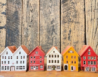 May include: Six miniature ceramic houses with red, white, and yellow paint. Each house has a different storefront with a name and a small sign. The houses are arranged in a row on a wooden surface.