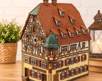 May include: A miniature model of a multi-story timber-framed building with a red tiled roof, dormer windows, and a green turret. The facade features yellow walls, brown shutters, and small flower boxes with orange blossoms. Text reads Bäckerei-Konditorei.