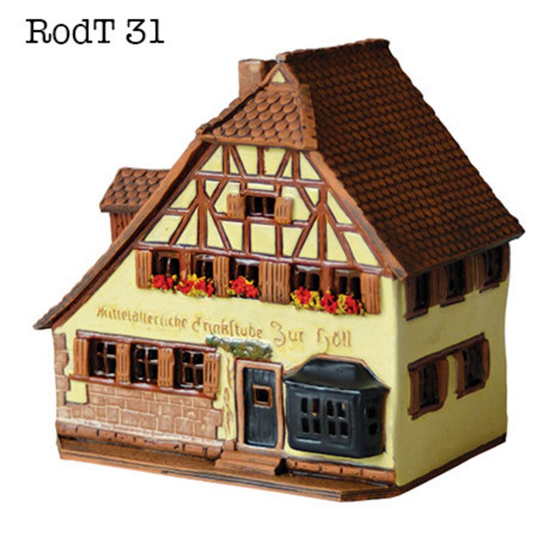 May include: A ceramic model of a traditional German half-timbered house with a brown tiled roof. The house has a sign that reads "Mittelalterliche Trinkstube Zur Holl".