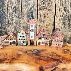 May include: Five miniature ceramic houses with intricate details, including a tall tower with a clock face, a green house with a sign that reads "Gasthaus Grun", a yellow house with a sign that reads "Zur Gaste", a white house with a sign that reads "B&auml;ckerei Braun", and a red house with a sign that reads "Gasthof zum Adler".