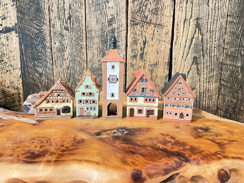 May include: Five miniature ceramic houses with intricate details, including a tall tower with a clock face, a green house with a sign that reads "Gasthaus Grun", a yellow house with a sign that reads "Zur Gaste", a white house with a sign that reads "B&auml;ckerei Braun", and a red house with a sign that reads "Gasthof zum Adler".