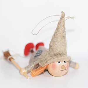 May include: A small, handmade witch doll with a brown felt hat, red shoes, and a wooden face. The doll is holding a small broom made of twigs.