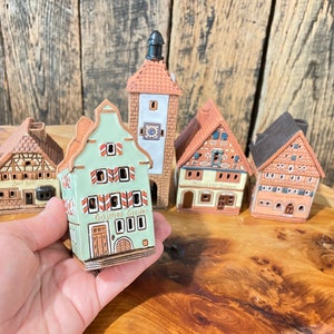 May include: Five miniature ceramic houses with intricate details. The houses are painted in shades of brown, green, and white. The houses have small windows and doors. One house has the text "Gasthof Greuter" on the front.