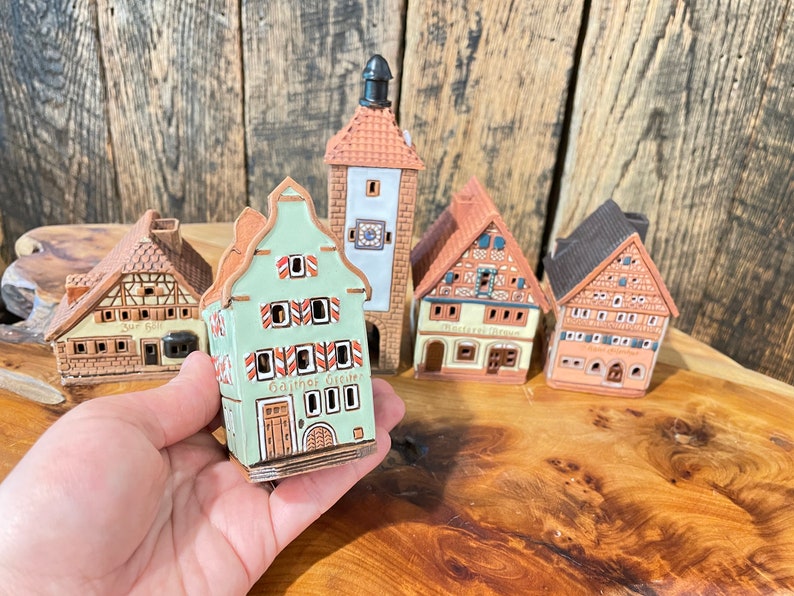 May include: Five miniature ceramic houses with intricate details. The houses are painted in shades of brown, green, and white. The houses have small windows and doors. One house has the text "Gasthof Greuter" on the front.
