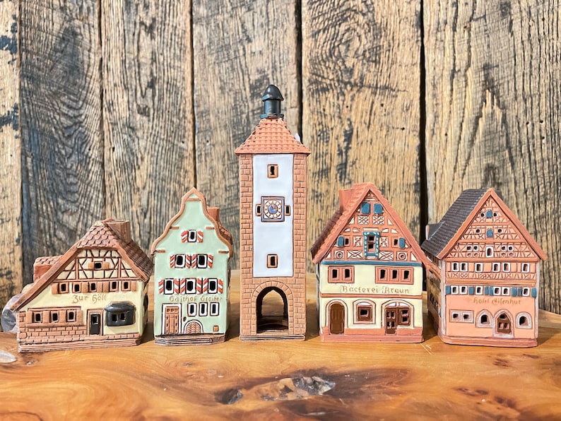 May include: Five miniature ceramic houses with intricate details, including a clock tower, a green house with the text "Gasthaus Greu", a brown house with the text "B&auml;ckerei Braun", and two other houses with German text.